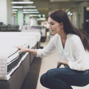 A woman in a white shirt and jeans in a mattress store. She examines the mattress she wants to buy. She squats and looks at the mattress